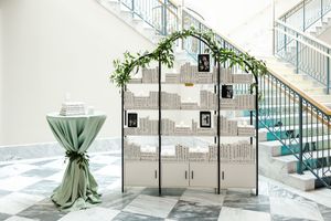 An elegant wedding seating chart display with sage green decor and metal archway in a bright modern venue.