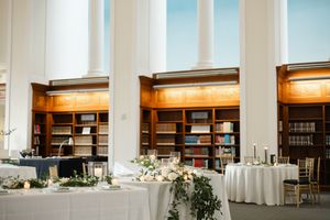 Elegant wedding reception tables with white linens and floral arrangements set against illuminated library bookshelves.