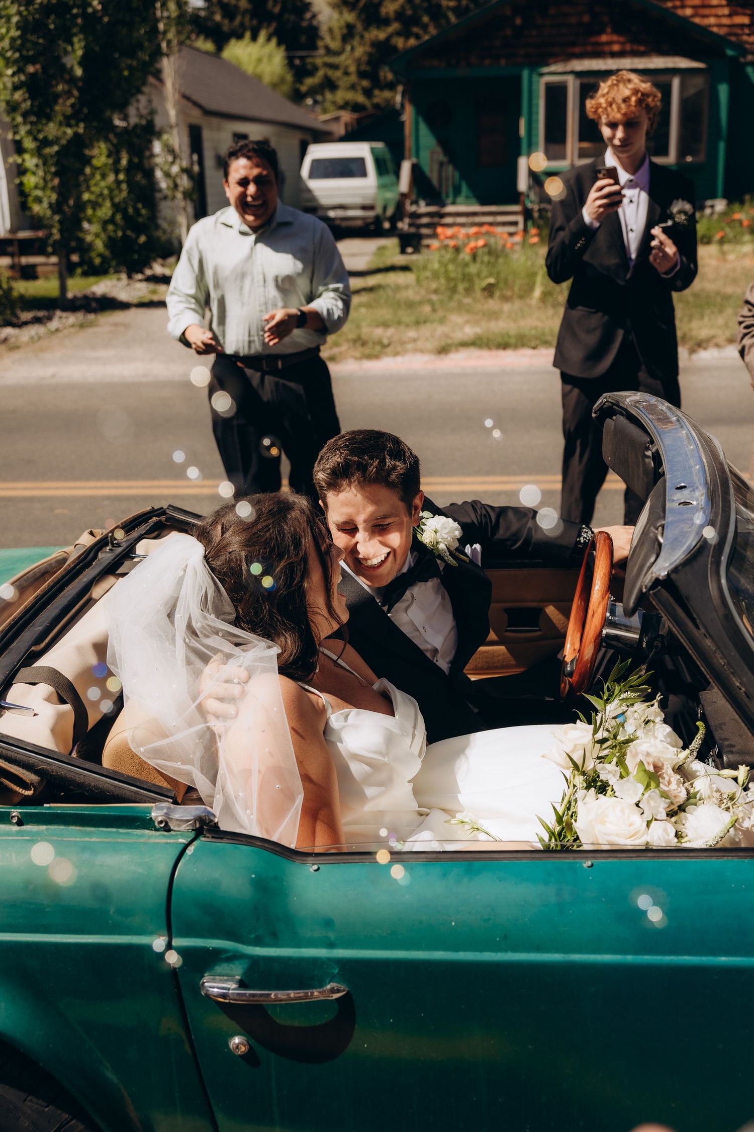 A happy couple sits in a vintage teal convertible car decorated with flowers and wedding attire on a sunny day.