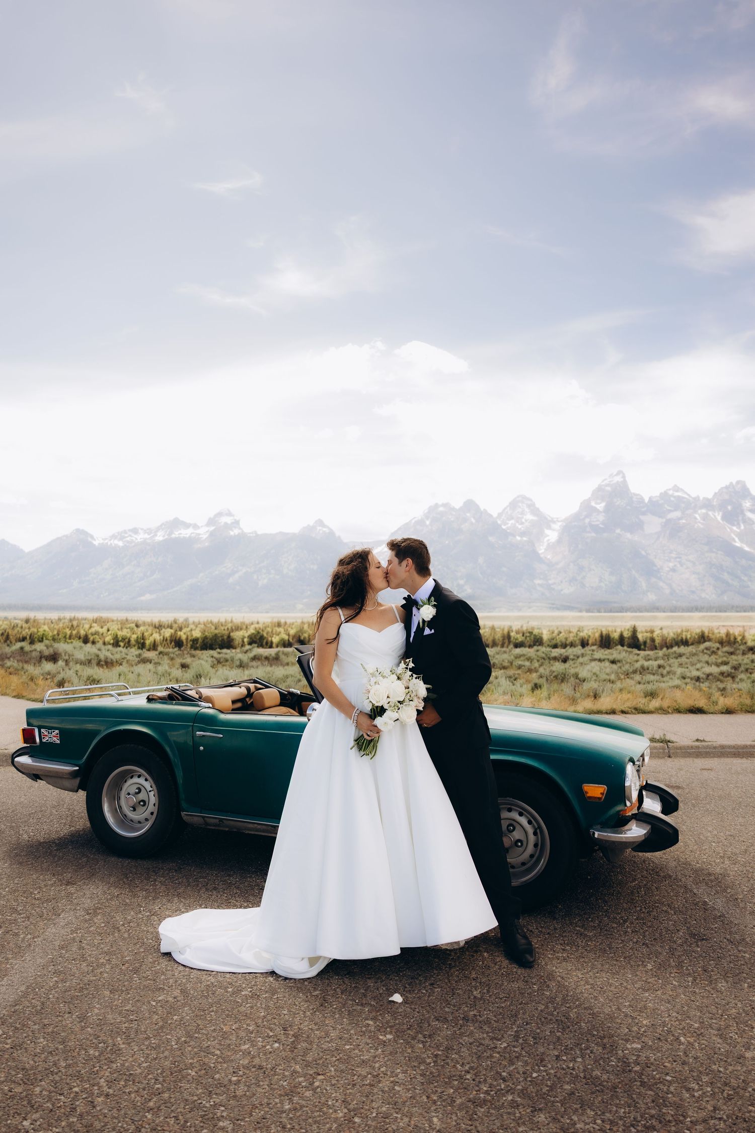 Couple shares romantic moment by vintage green convertible with Grand Teton mountains in background.