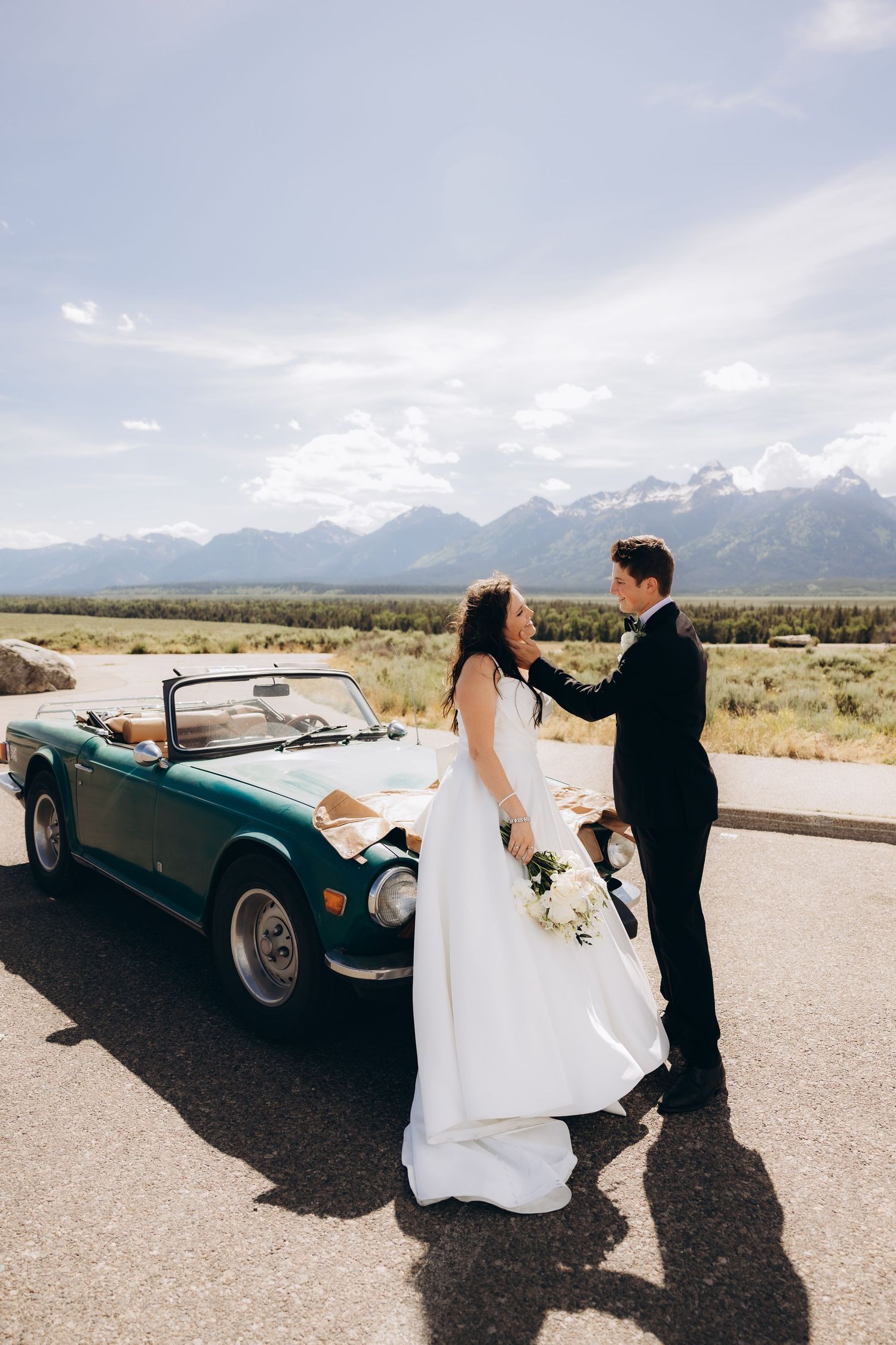 Wedding couple stands beside vintage green convertible car with majestic mountain range in background.
