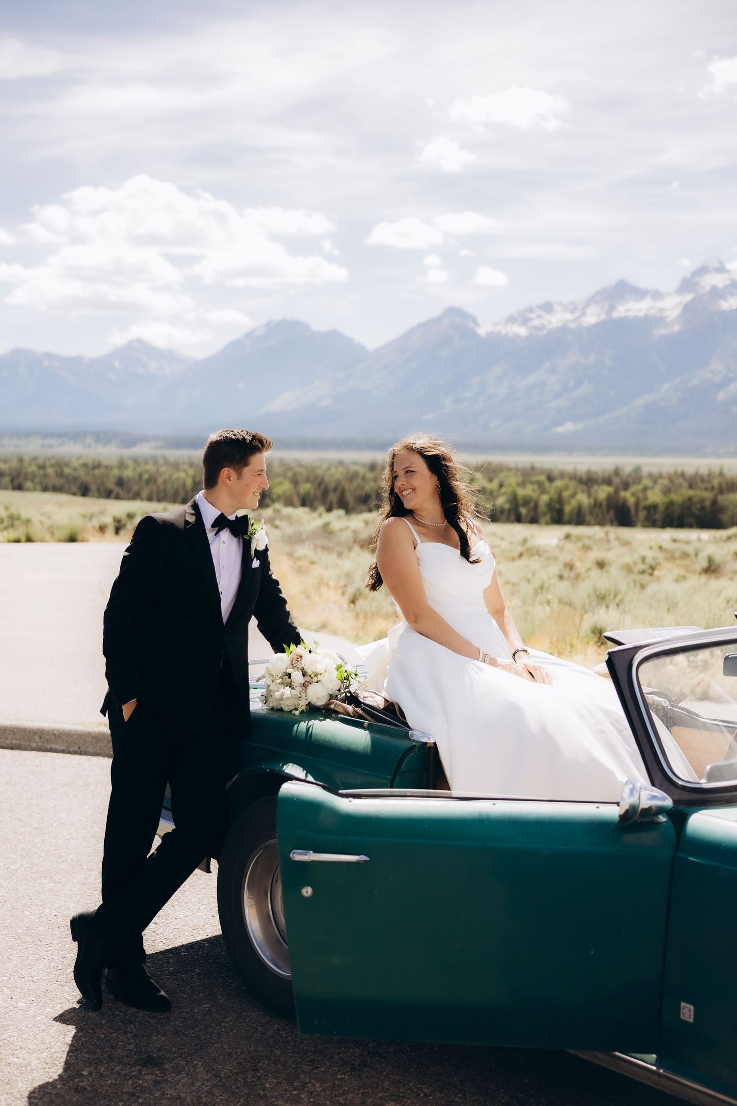 A bride and groom share an intimate moment beside a classic vintage car with scenic mountains in the background.