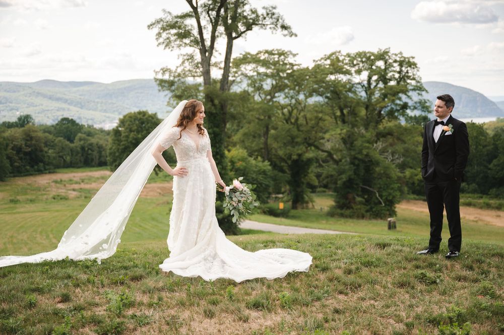 Couple stands in rolling green meadow with flowing white dress and veil creating romantic wedding portraits in nature.