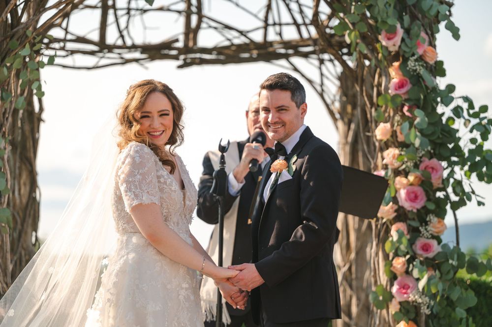 A joyful wedding ceremony takes place under a rustic wooden arch decorated with pink roses and greenery.