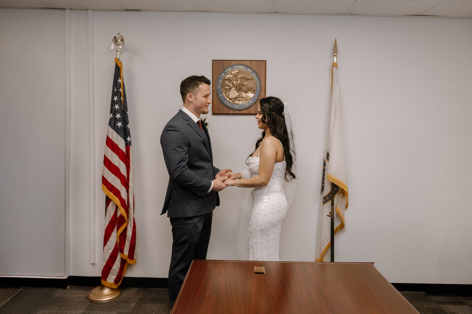 A couple celebrates their wedding ceremony in front of an American flag in a simple indoor venue.