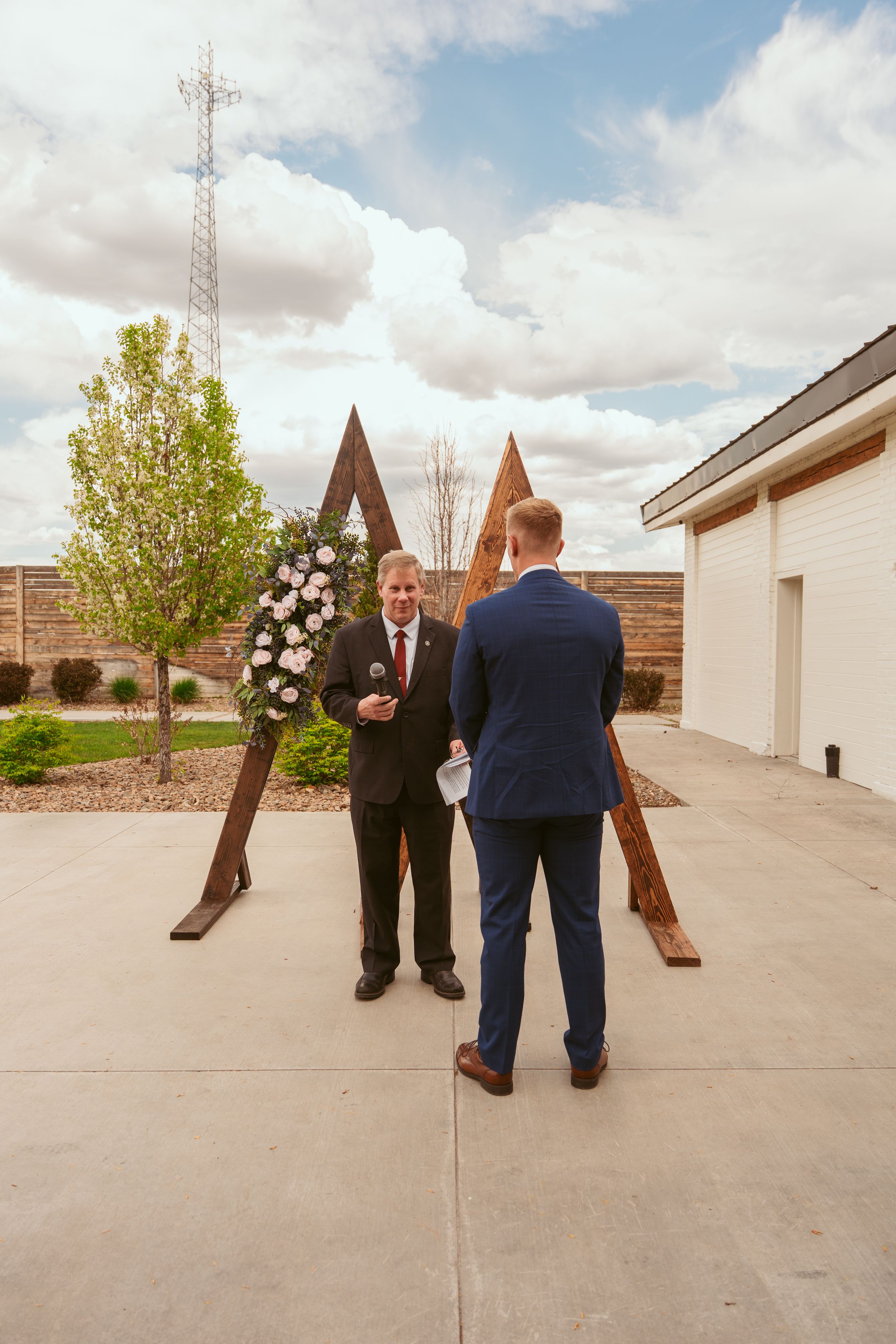 A contemporary wooden arch decorated with white flowers stands in an outdoor ceremony space with two people beneath it.