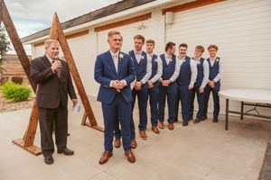 Wedding party members in navy suits and vests pose by a rustic wooden structure on a patio.