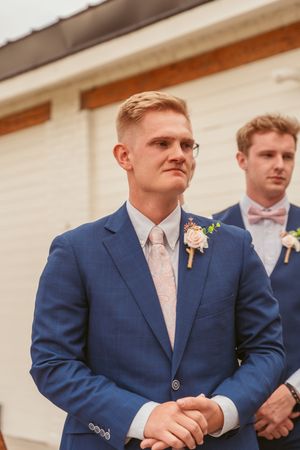 Wedding attendee in a navy blue suit with glasses and boutonniere stands outside during ceremony.