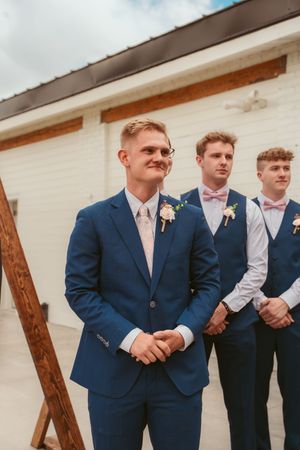Row of groomsmen in navy blue suits and pink ties stand together at a wedding ceremony.
