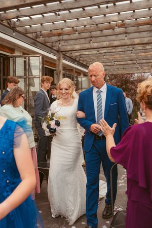 Wedding party mingles and laughs together under a rustic wooden pergola with natural lighting at an outdoor venue.