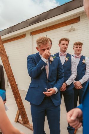 Emotional moment at wedding ceremony with groomsmen in matching navy blue suits standing near white wall and wooden beam.