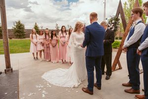 Wedding ceremony taking place outdoors with bridesmaids in pink dresses watching the couple exchange vows under wooden posts.