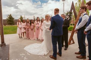 An outdoor wedding ceremony taking place under a wooden arch with attendants in pink dresses and brown suits.