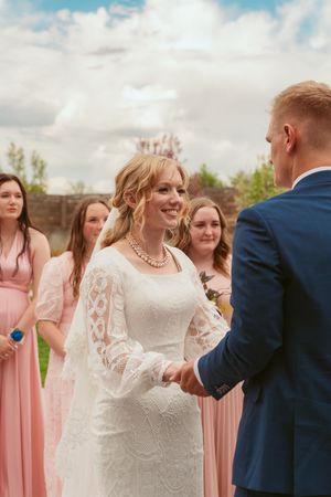 Wedding ceremony outdoors with bride in lace dress and groom in navy suit surrounded by bridesmaids in pink dresses.