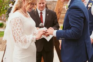 Close up view of couple exchanging rings during wedding ceremony with officiant in center wearing red tie.