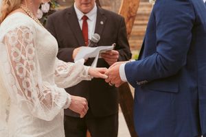 Close-up of hands exchanging wedding rings during ceremony with officiant in red tie standing behind couple.