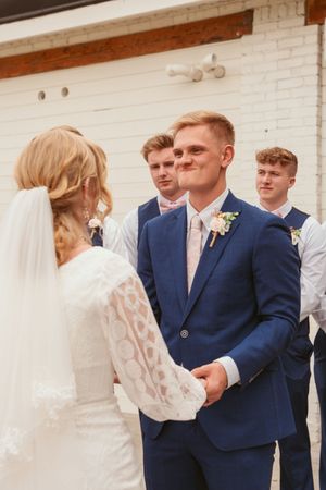 A wedding receiving line with navy-suited groomsmen and bridesmaids in pink dresses greeting guests in front of a white wall.