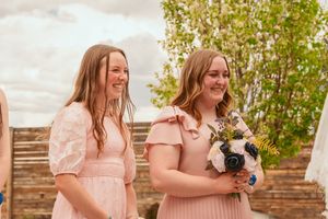 Two bridesmaids wearing blush pink dresses stand together holding bouquets during an outdoor wedding ceremony.