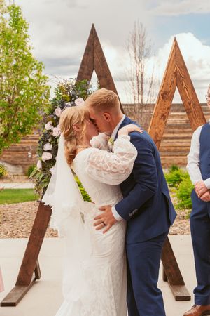 A couple shares their first kiss as newlyweds in front of rustic wooden triangular structures at an outdoor wedding venue.