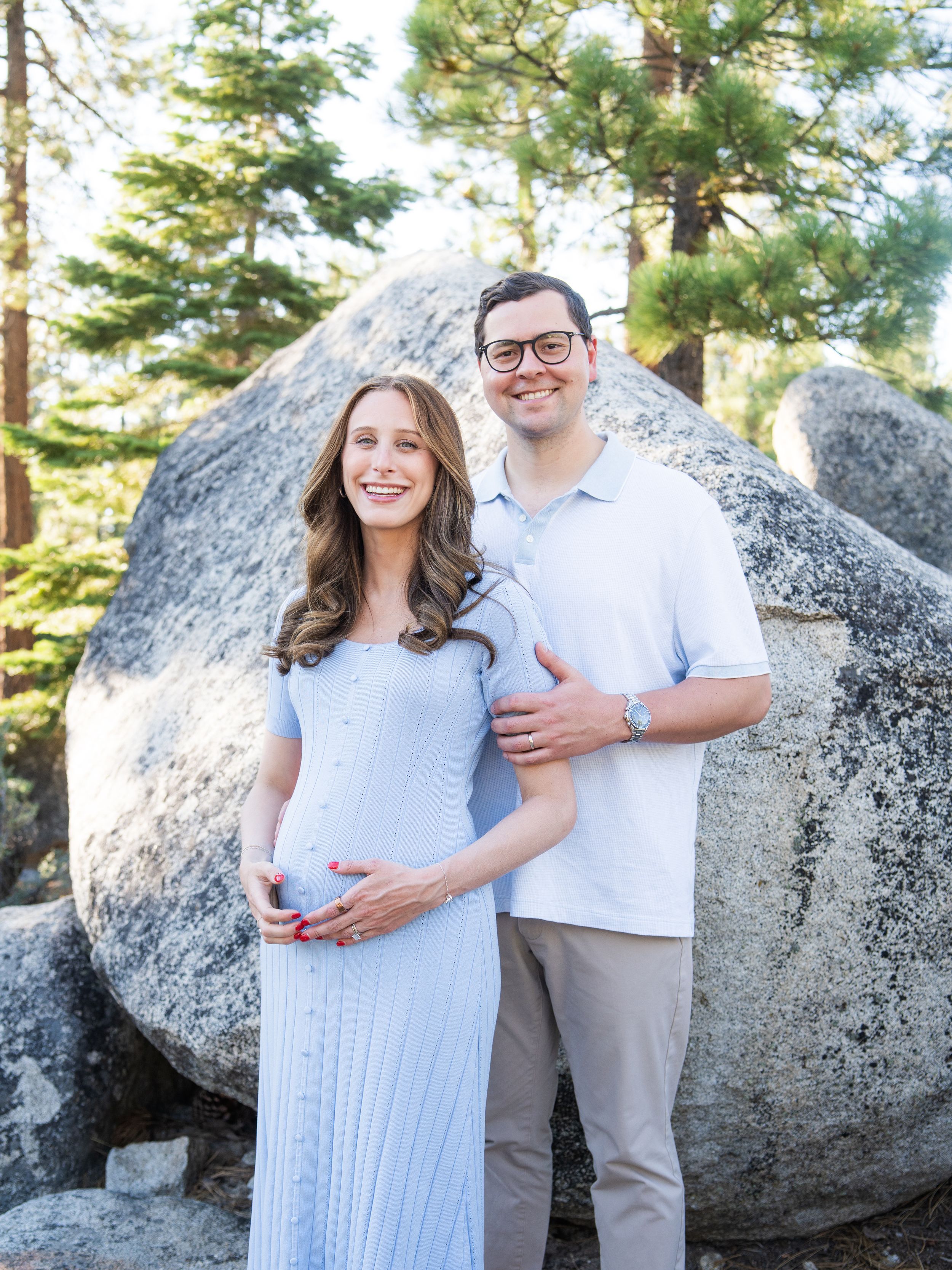 A couple poses together in front of large granite boulders and tall pine trees during a maternity photo session outdoors.