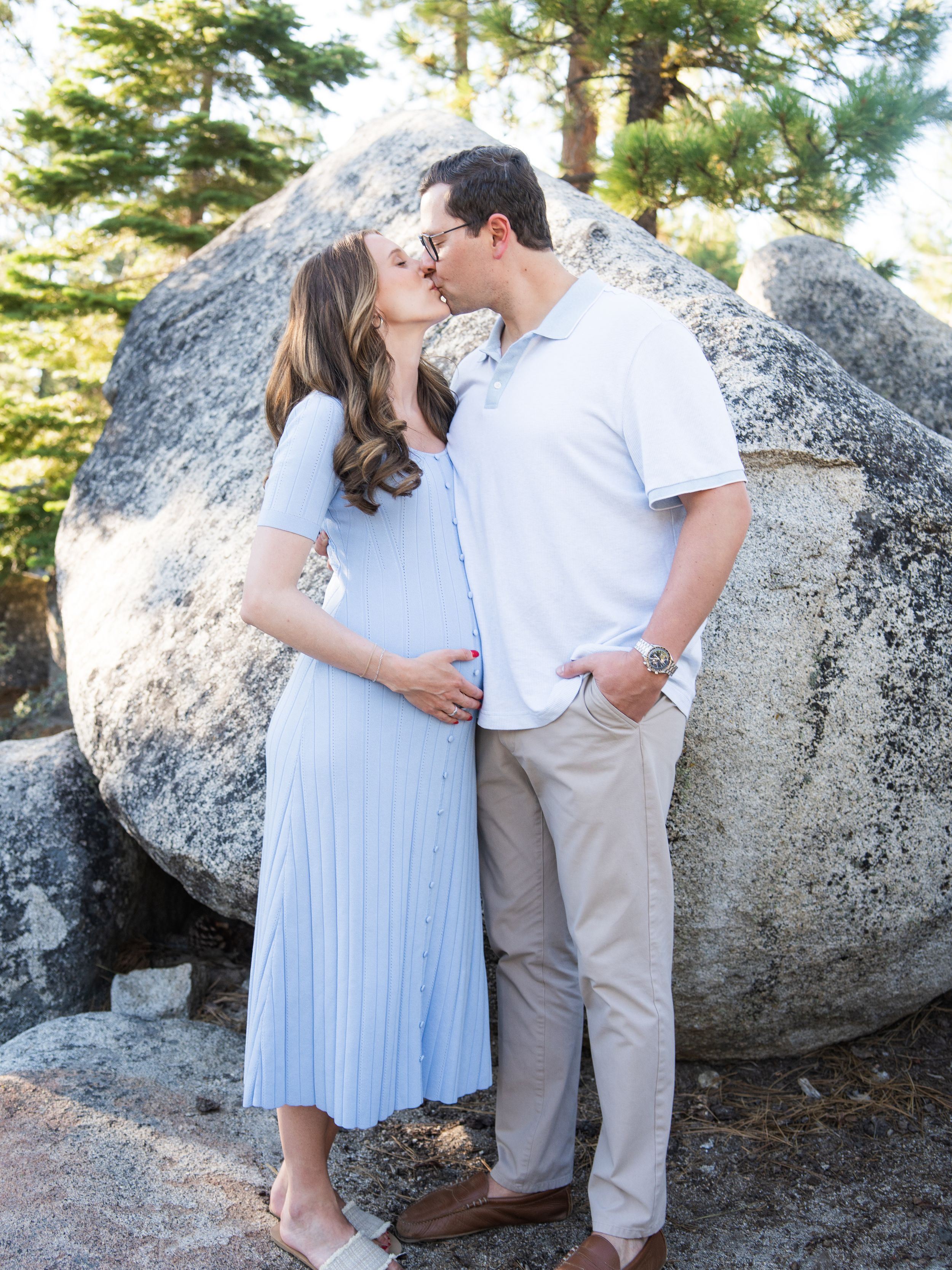 A romantic maternity moment captured in nature with a couple sharing a kiss against large granite boulders and pine trees.