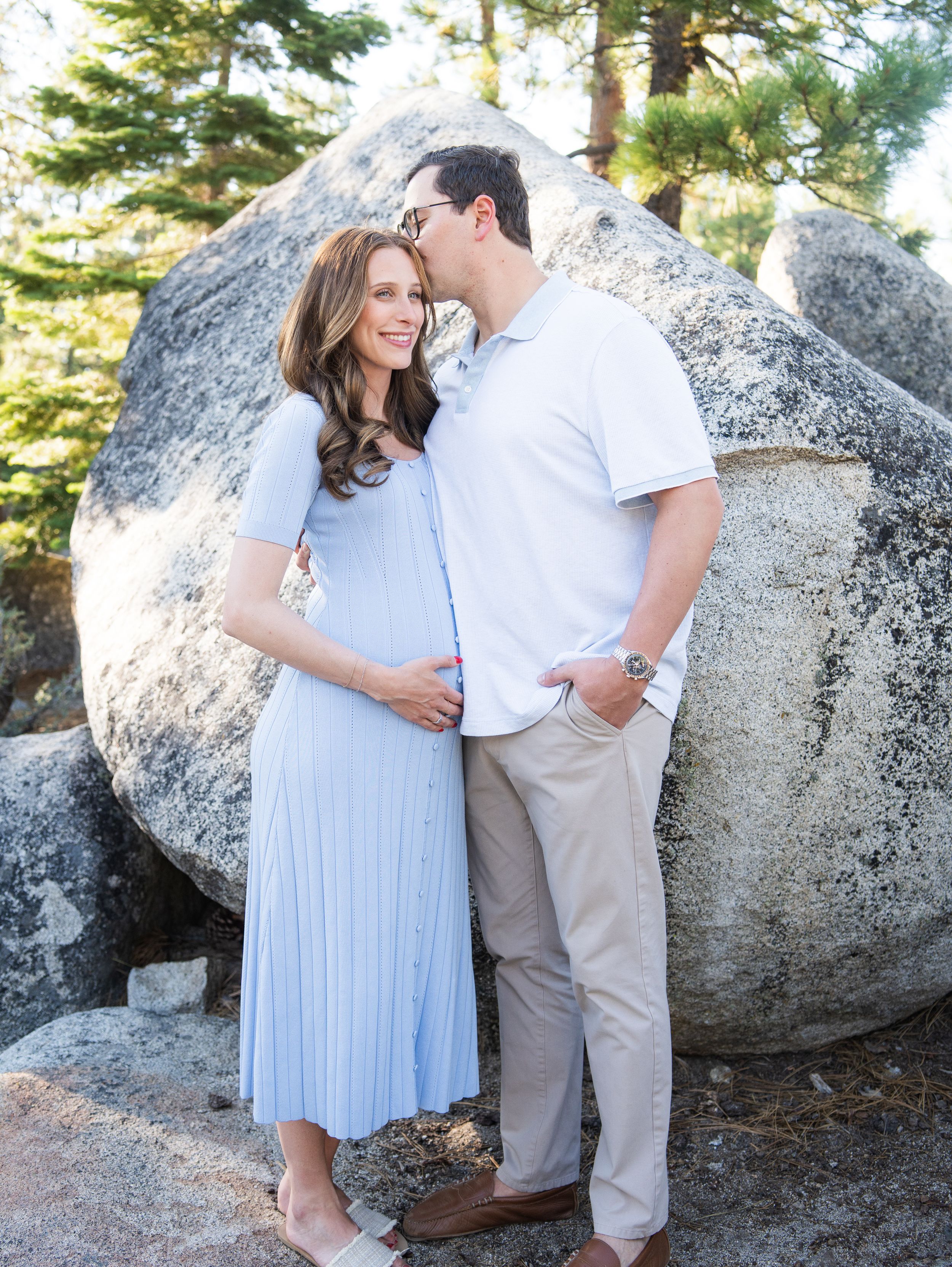 Couple in light blue dress and white shirt share an intimate moment against large granite boulders in a forest setting.
