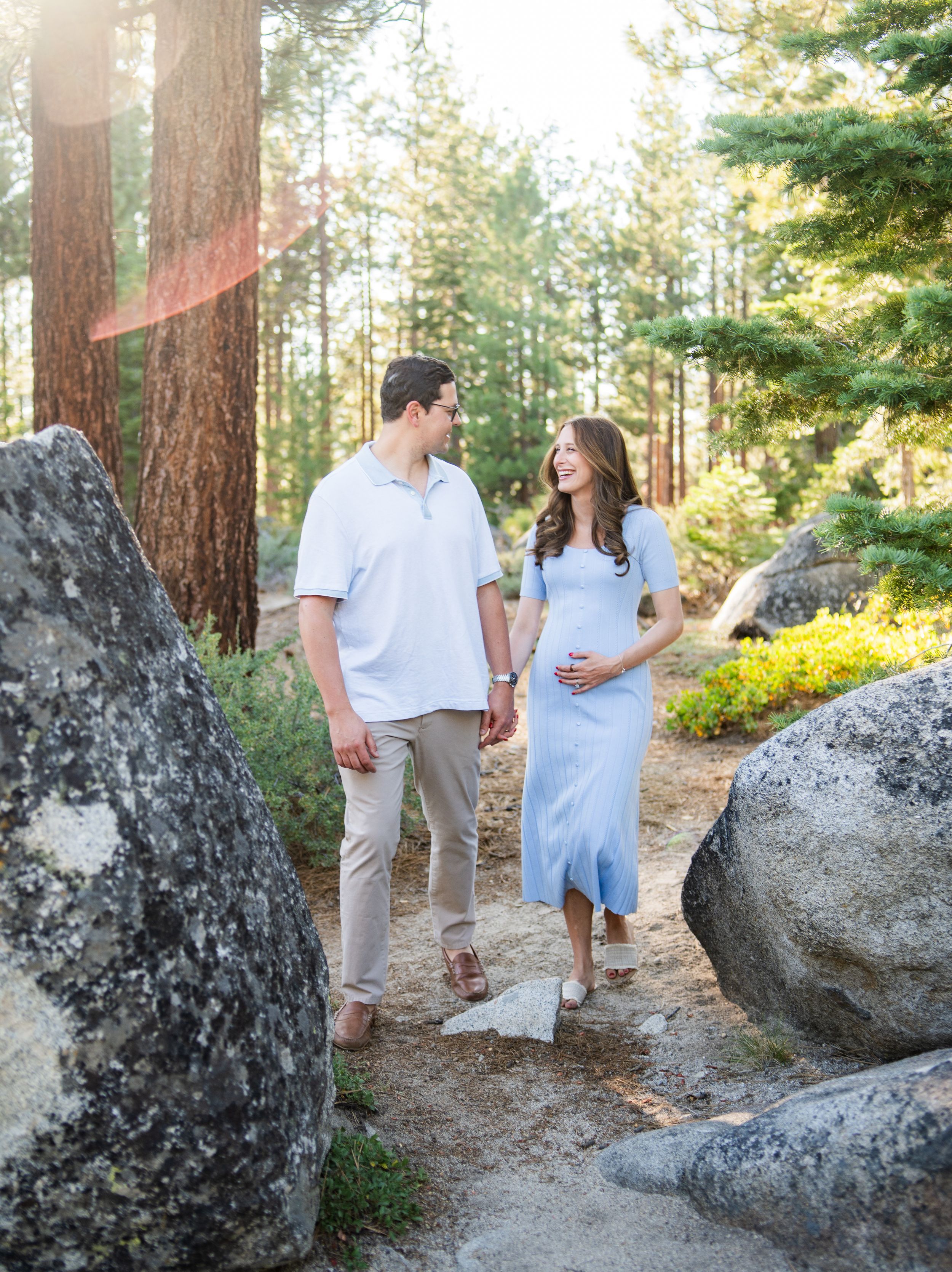 A couple walks hand in hand on a forest path between large boulders as sunlight filters through tall evergreen trees.