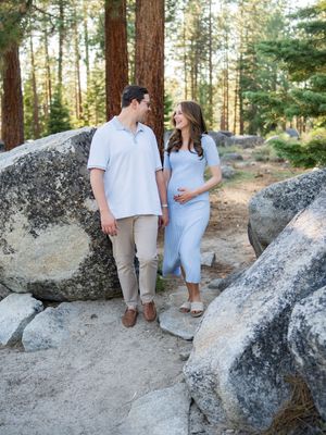 A couple stands together among large boulders in a pine forest, sharing a sweet moment in natural lighting.