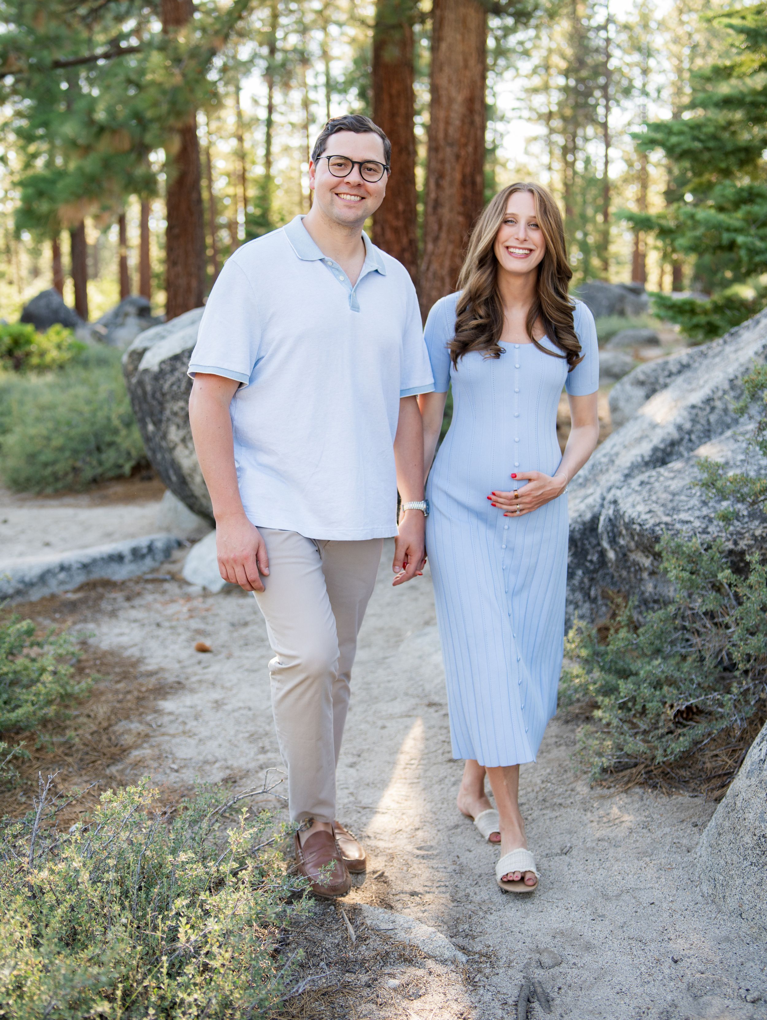 A couple in casual attire walks along a sunlit forest path during a romantic engagement photoshoot in nature.
