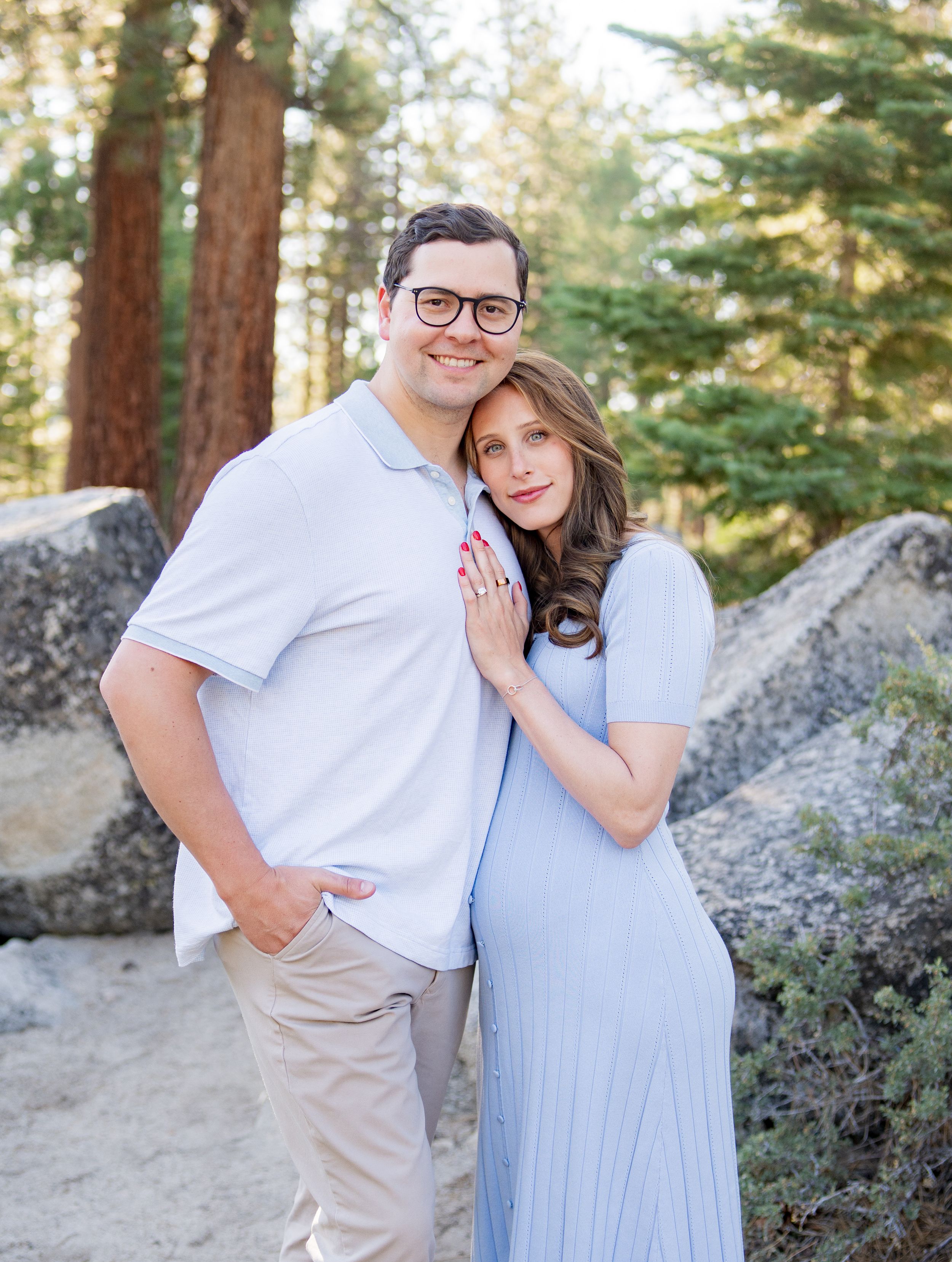 Couple in white and blue outfits embracing while standing against large boulders in a forest setting.