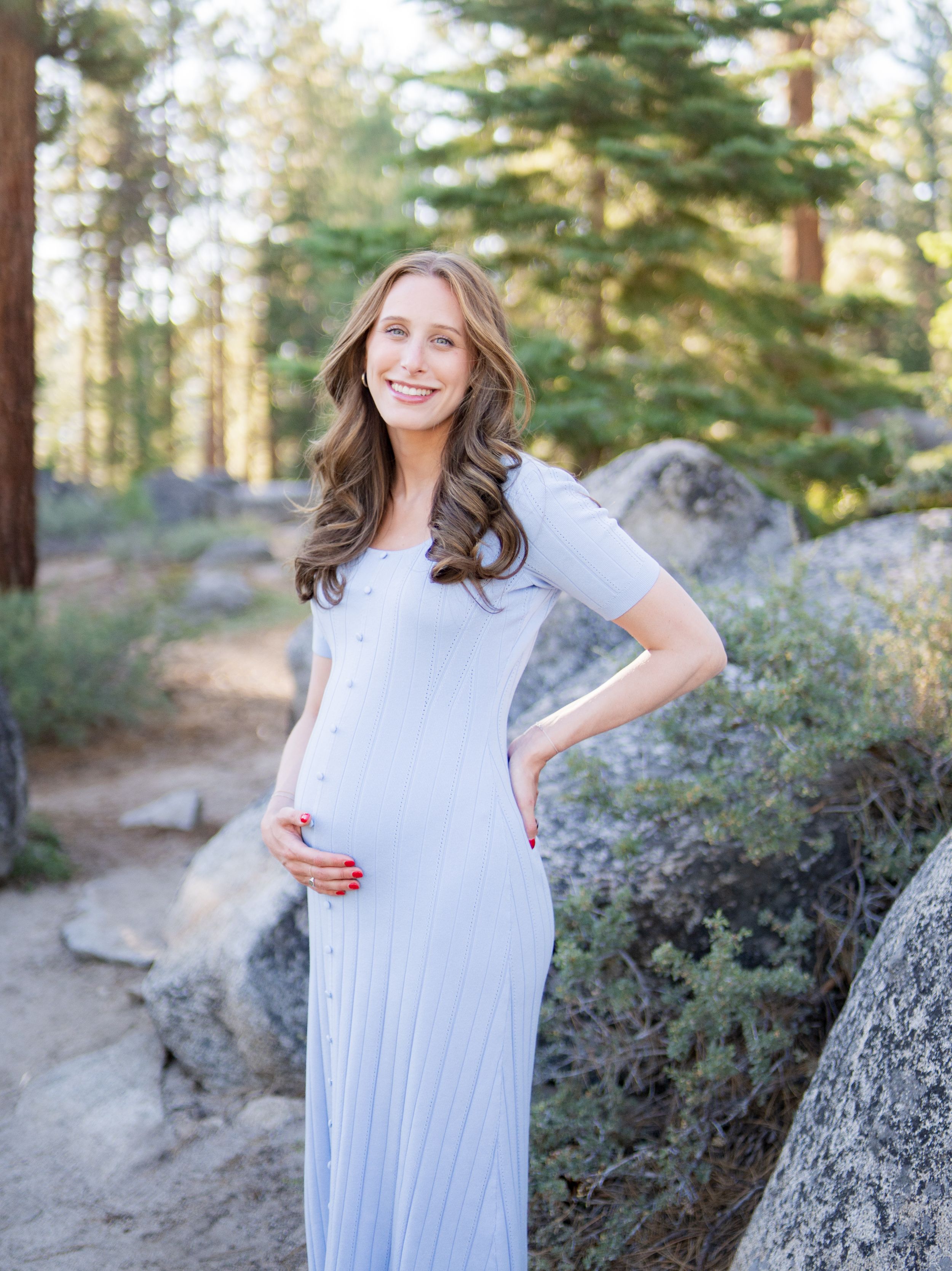 A person wearing a light blue dress poses outdoors in a wooded area with rocks and greenery visible in the background.