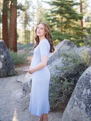 A person in a flowing pale blue dress poses on a forest path surrounded by large boulders and tall pine trees.