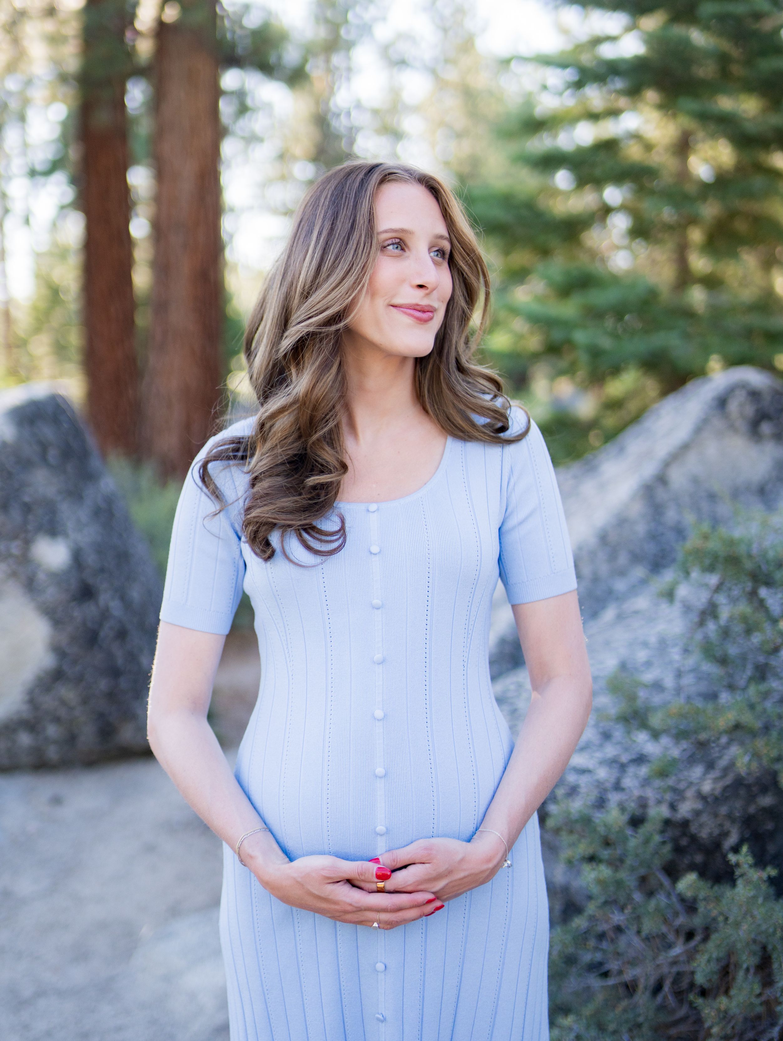 Woman in light blue dress stands in sunlit forest setting with evergreen trees and boulders in background.