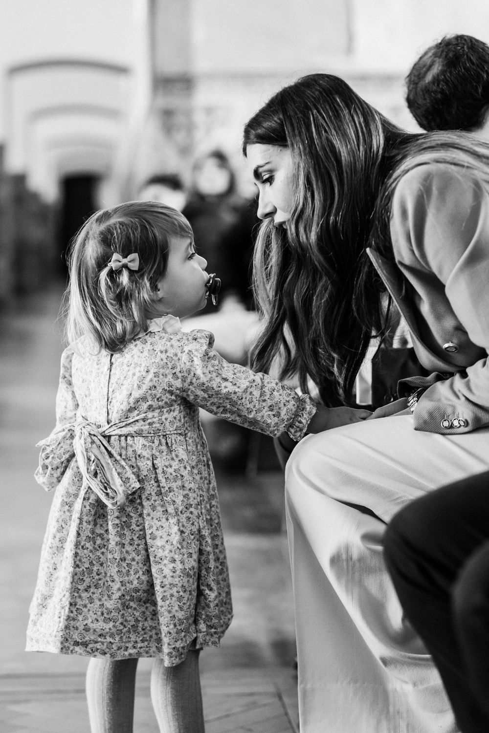 A young girl in a floral dress with bow looks up while interacting with someone seated on a bench in black and white.