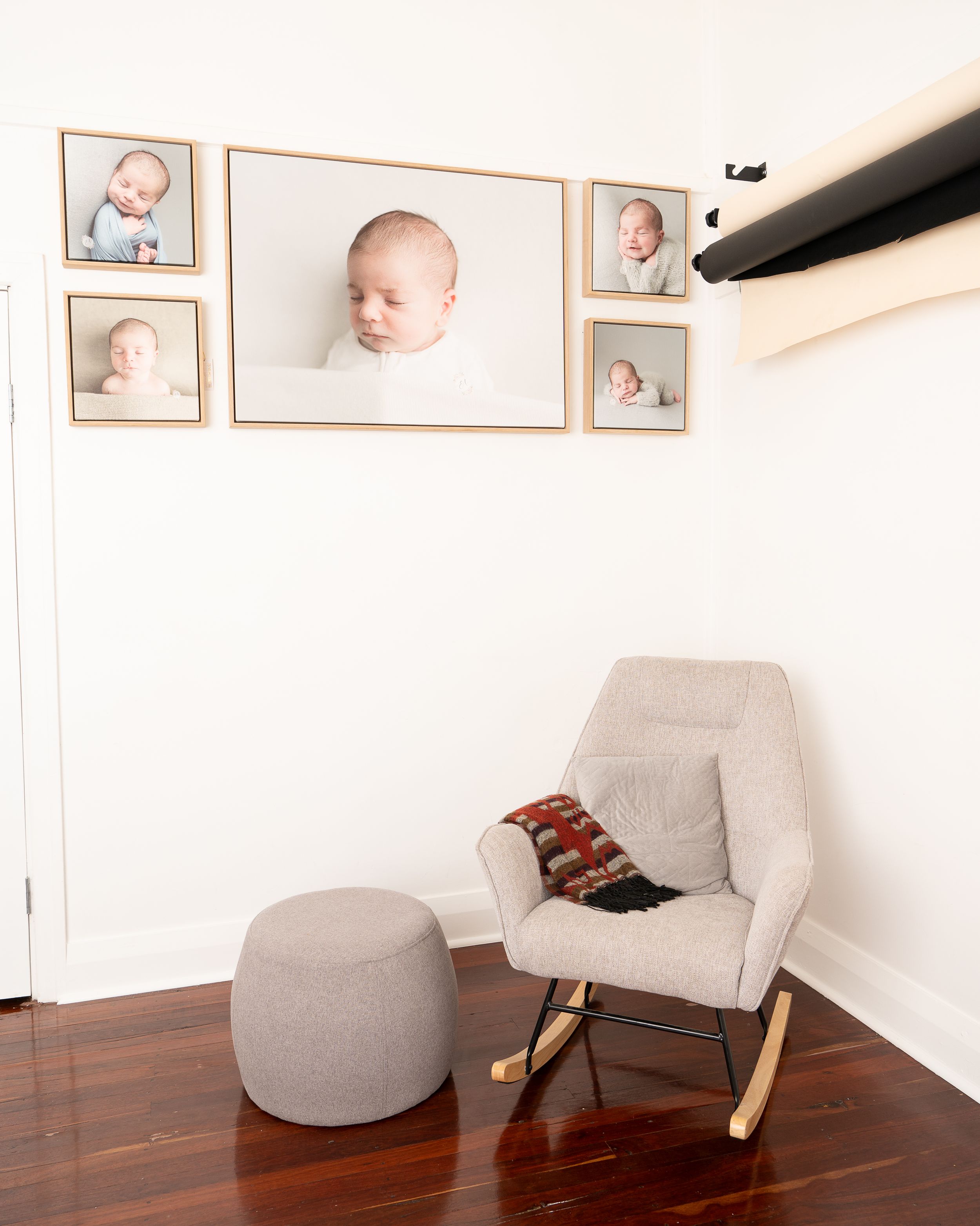 Cozy nursery corner with a gray rocking chair, ottoman and framed baby photos on white walls.