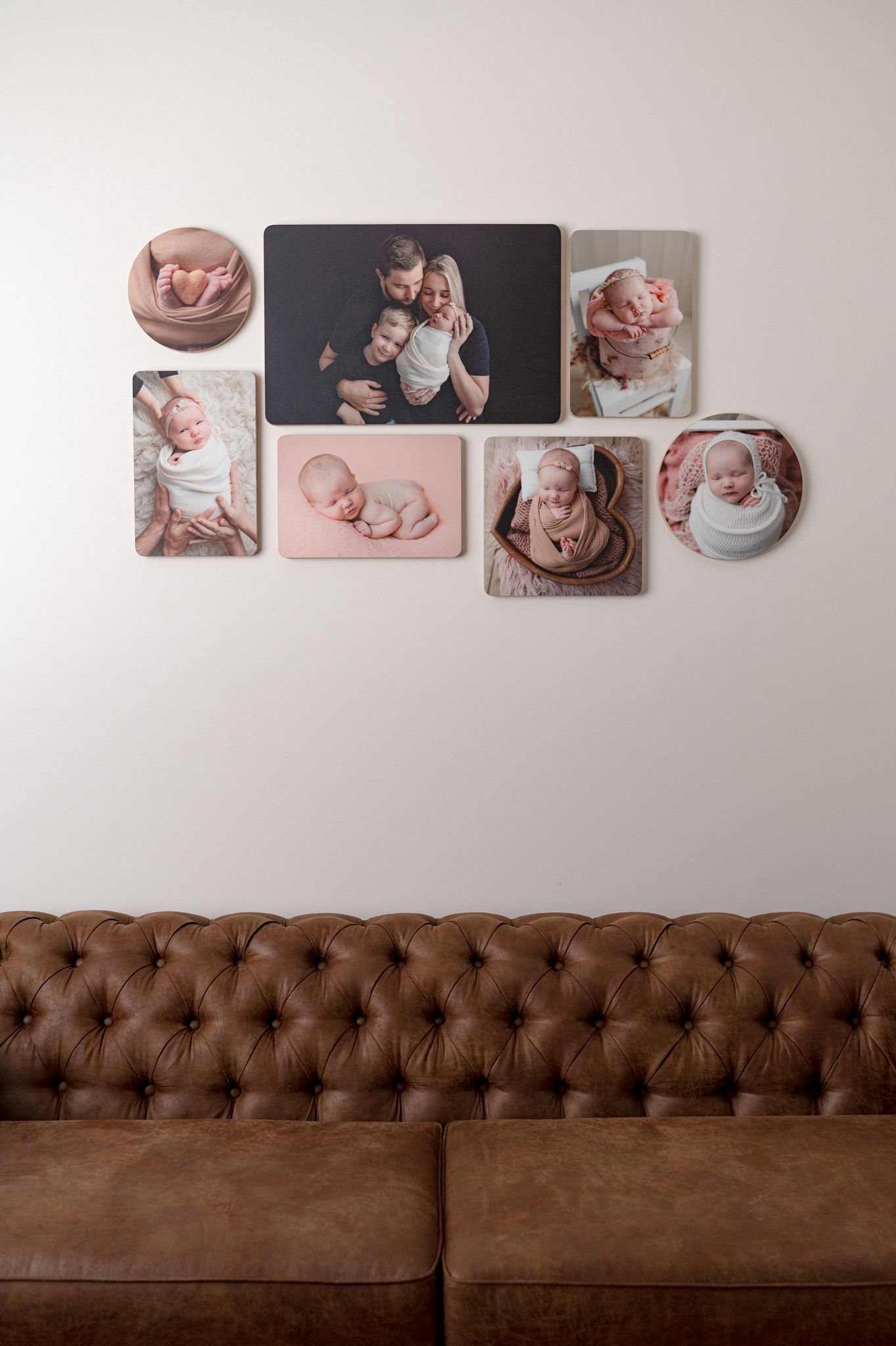 A collection of framed baby photos displayed above a brown tufted leather sofa against a white wall.
