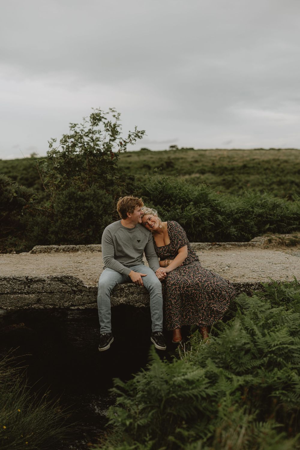 A romantic couple sits together on rocks surrounded by lush greenery in moody overcast lighting.