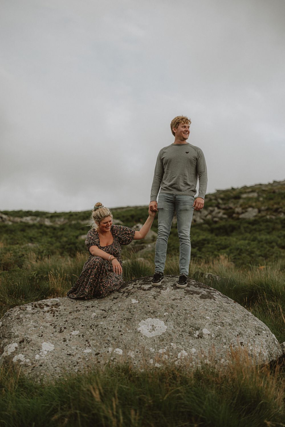 A couple sits together on a large boulder surrounded by moorland vegetation.