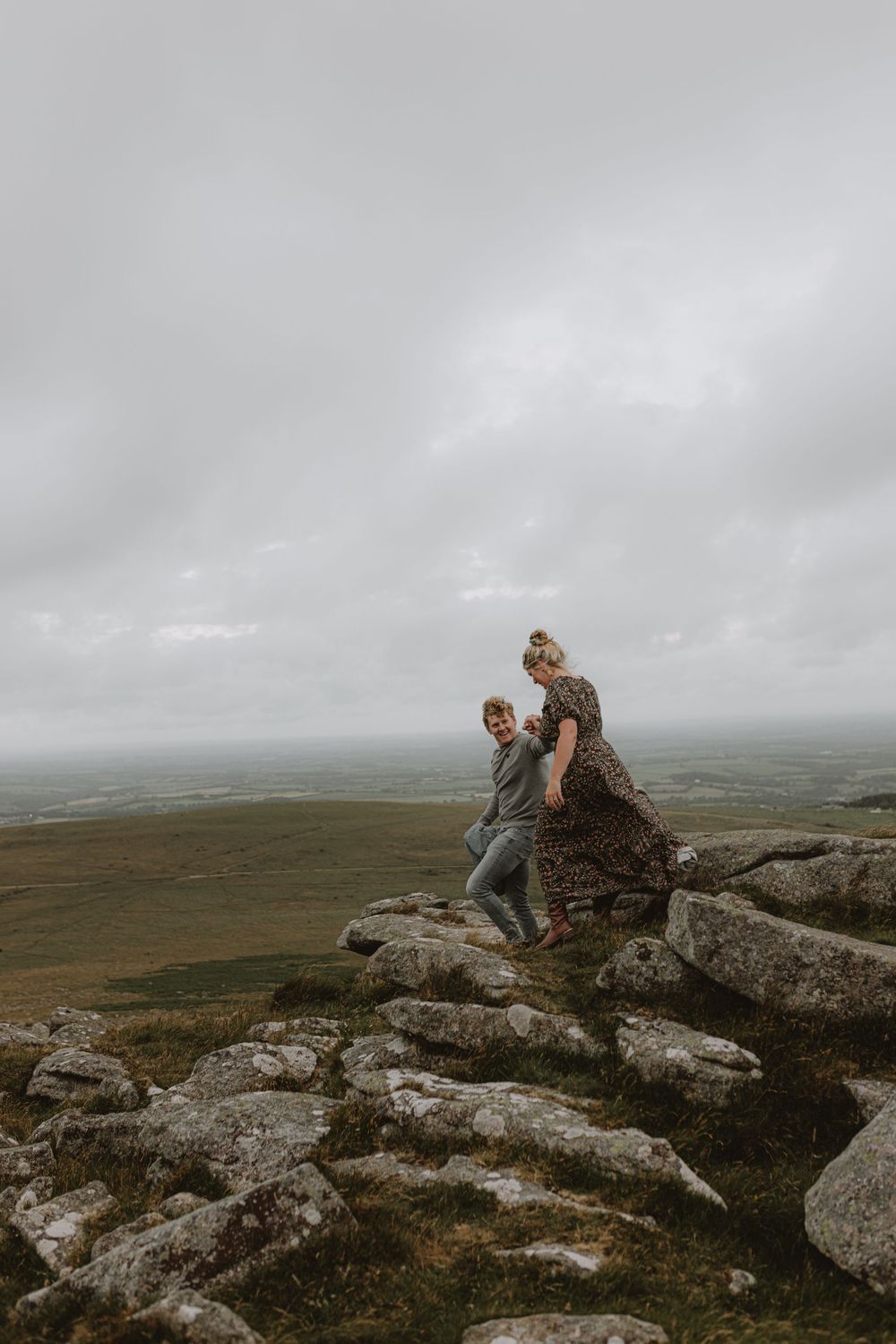 A couple rests on rocky outcrops overlooking a vast moorland landscape.
