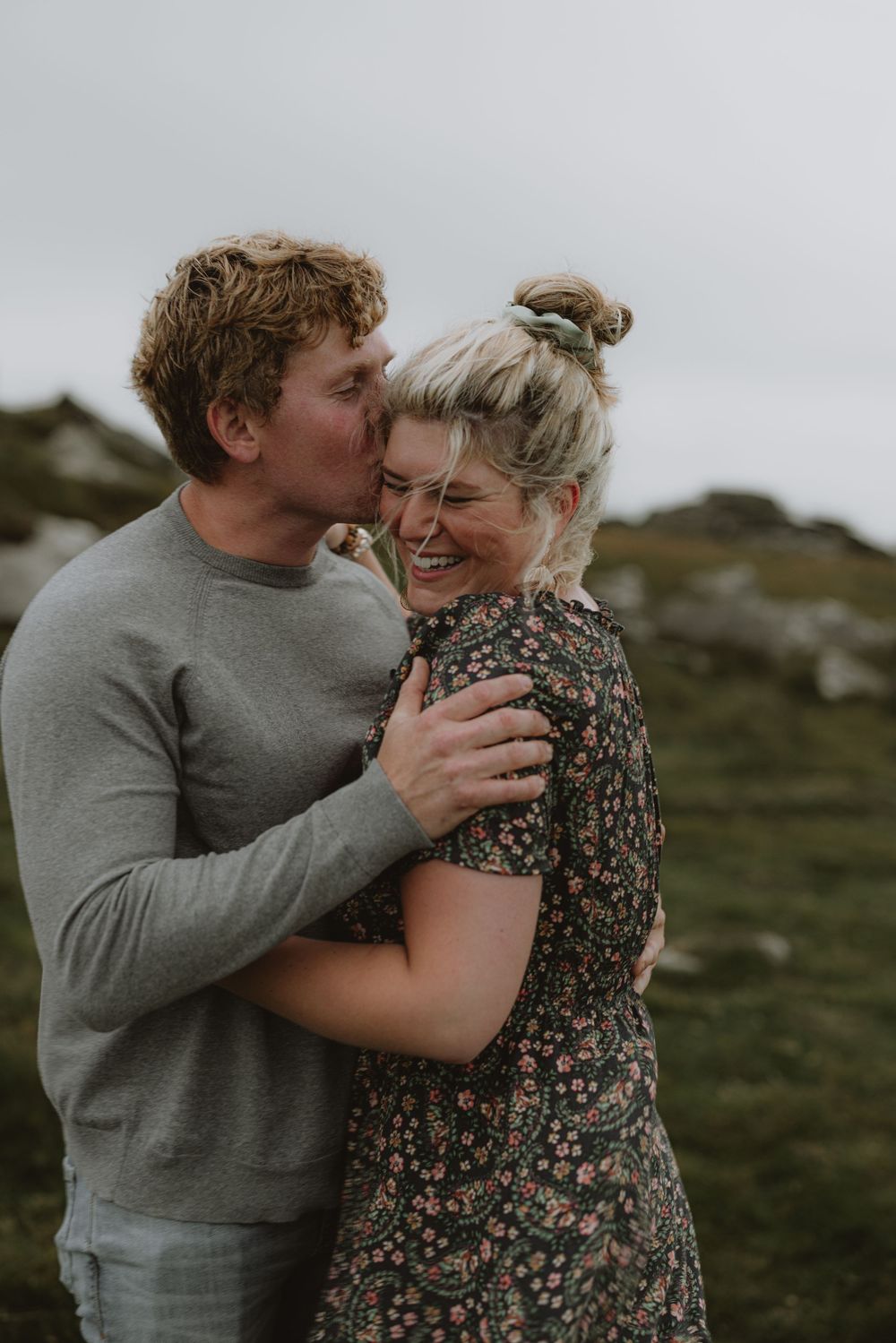 A romantic couple embraces and laughs together on a windswept hilltop with moody grey skies in the background.