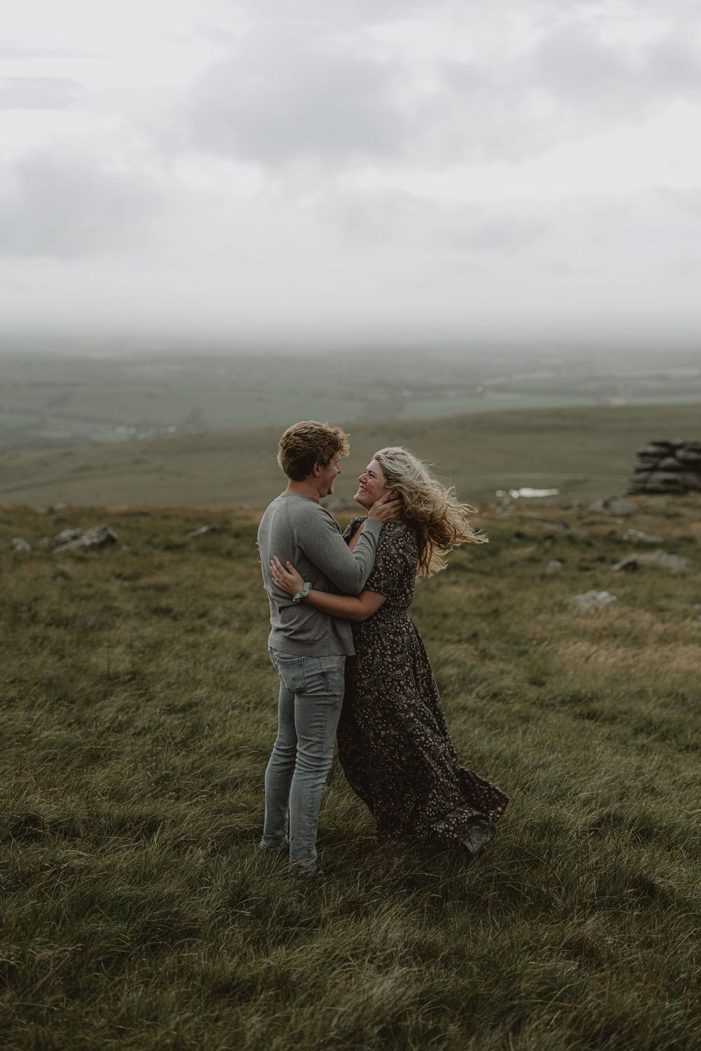 A couple shares an intimate moment while standing in tall grass on a misty hillside.