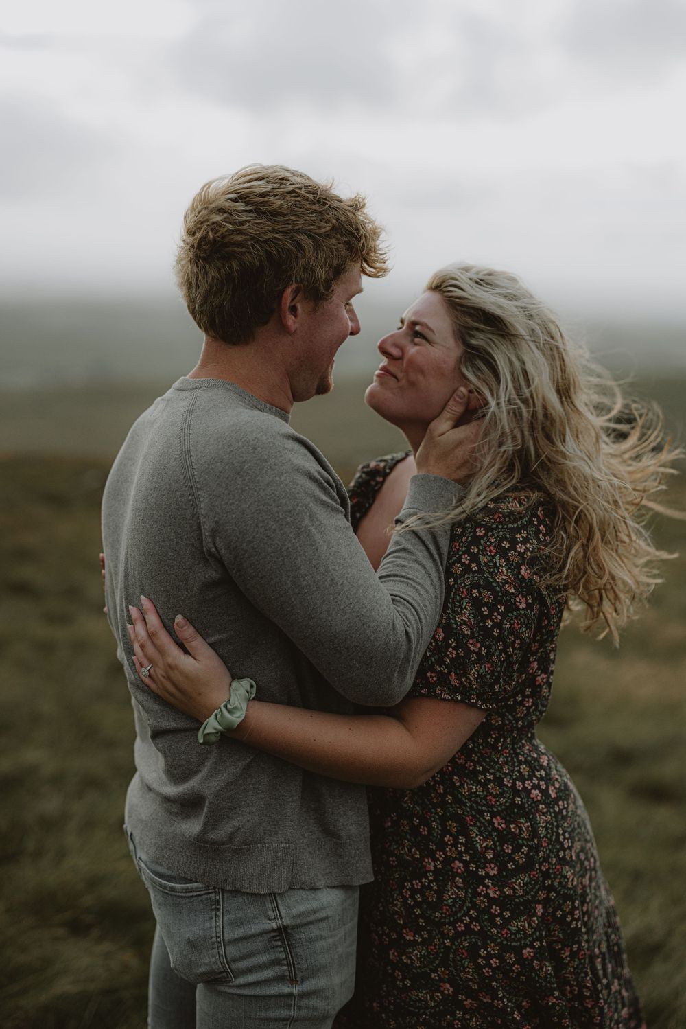 Two people share joyful laughter while embracing on a windy hilltop with dramatic cloudy skies.