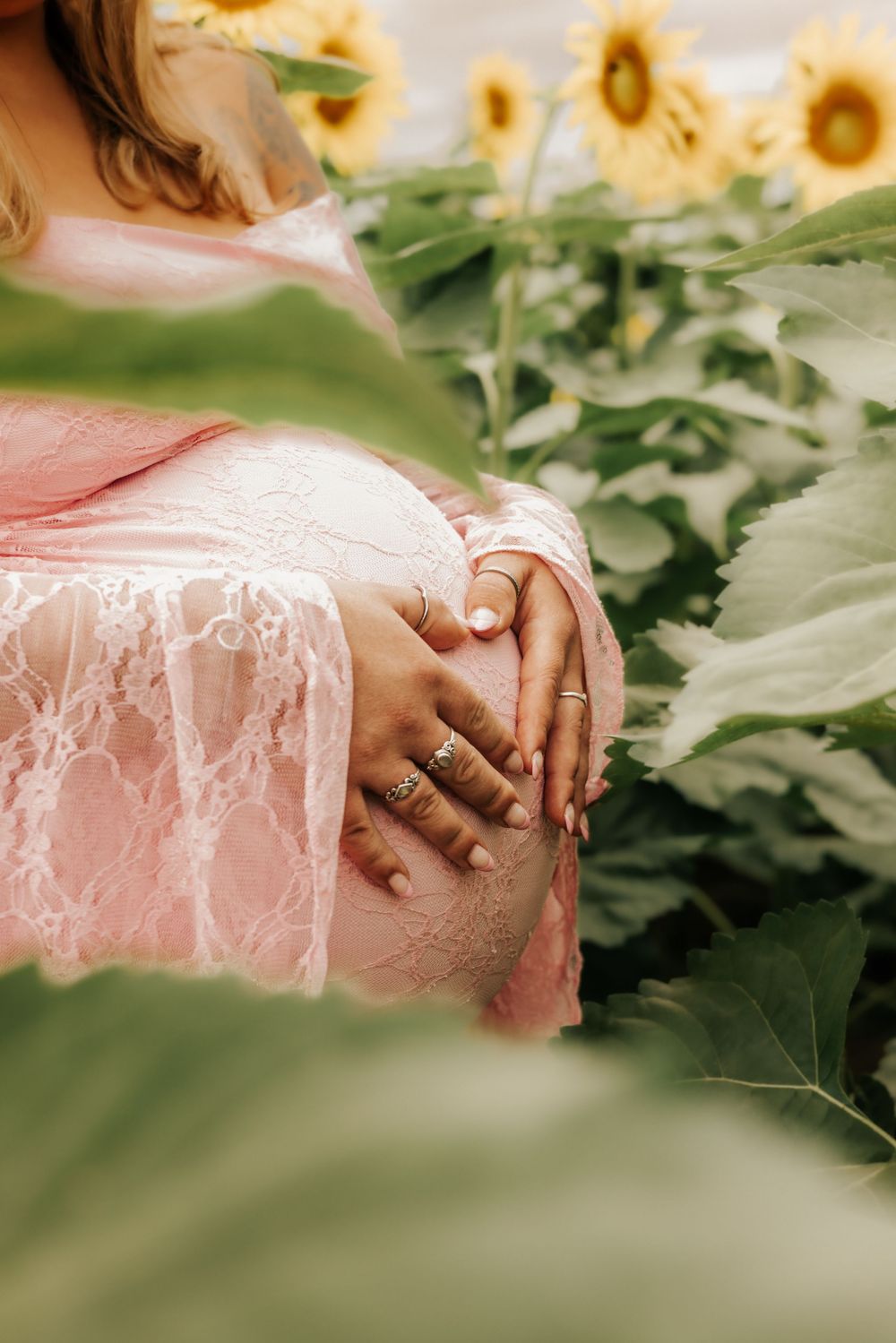 Close-up photo of hands with engagement rings amid sunflowers and greenery wearing a pink lace dress.
