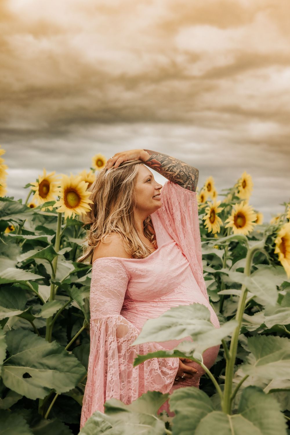 A dreamy portrait in a sunflower field features a person wearing a pink off-shoulder dress against moody cloudy skies.