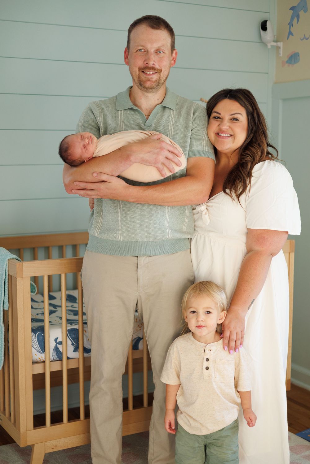 Happy family of four posing in nursery, parents holding newborn baby with toddler son standing in front.