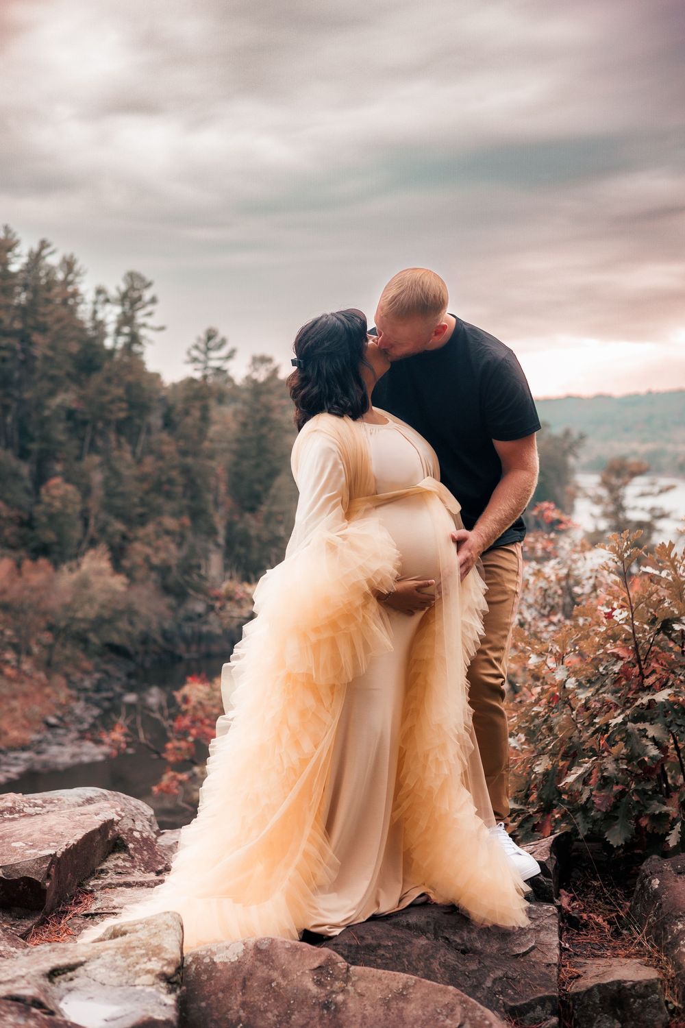 A romantic couple in formal attire poses together on rocky cliffs surrounded by autumn foliage at sunset.