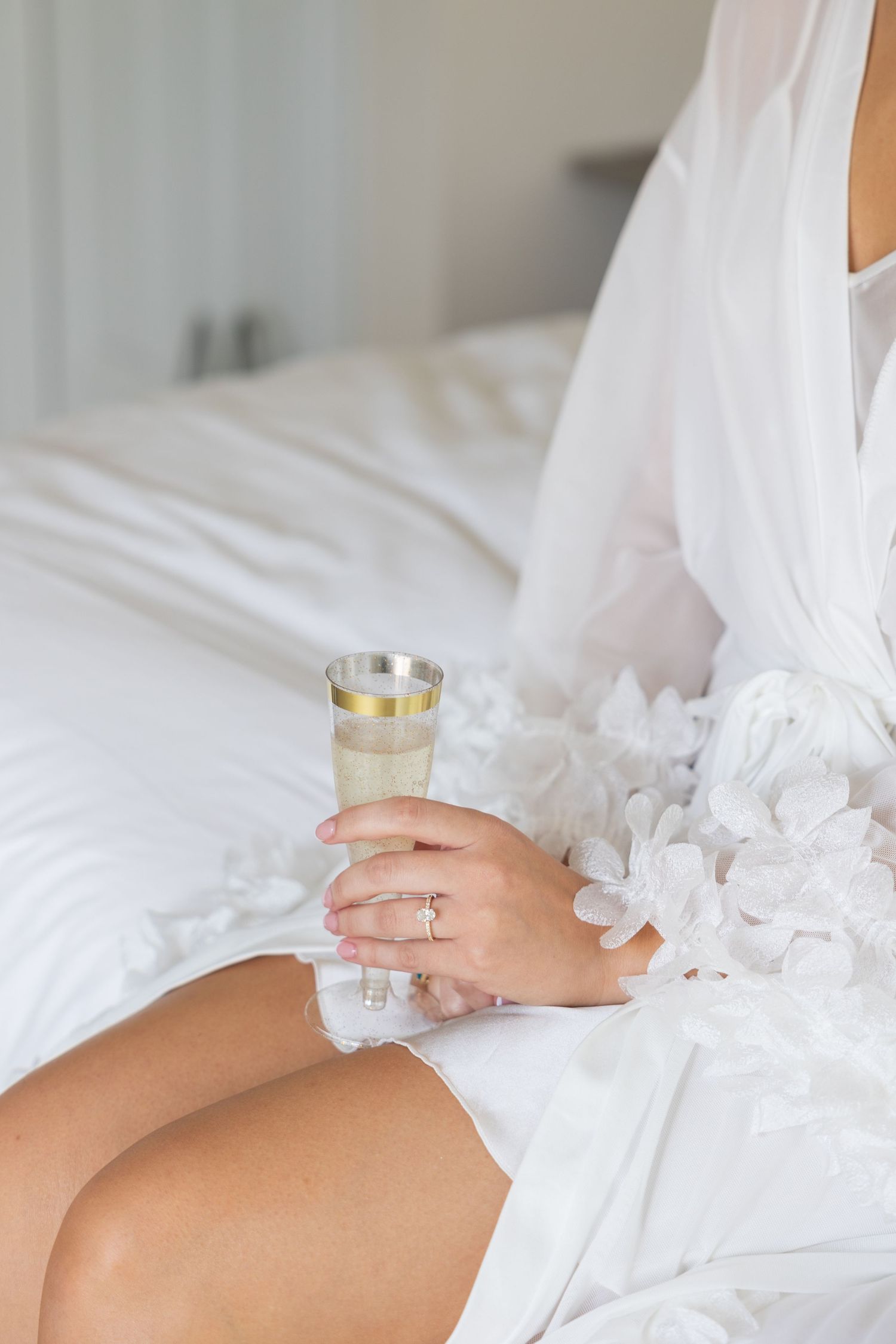 Person in white robe holding champagne glass while sitting on bed with engagement ring visible on hand. Rachel Martinez Photography