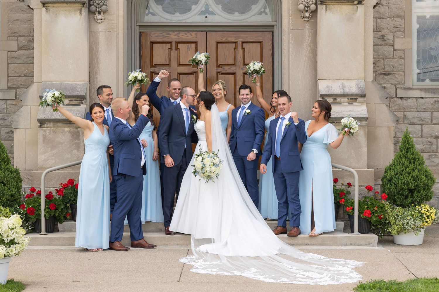 Wedding party in navy suits and light blue dresses poses on stone church steps with elegant floral bouquets.