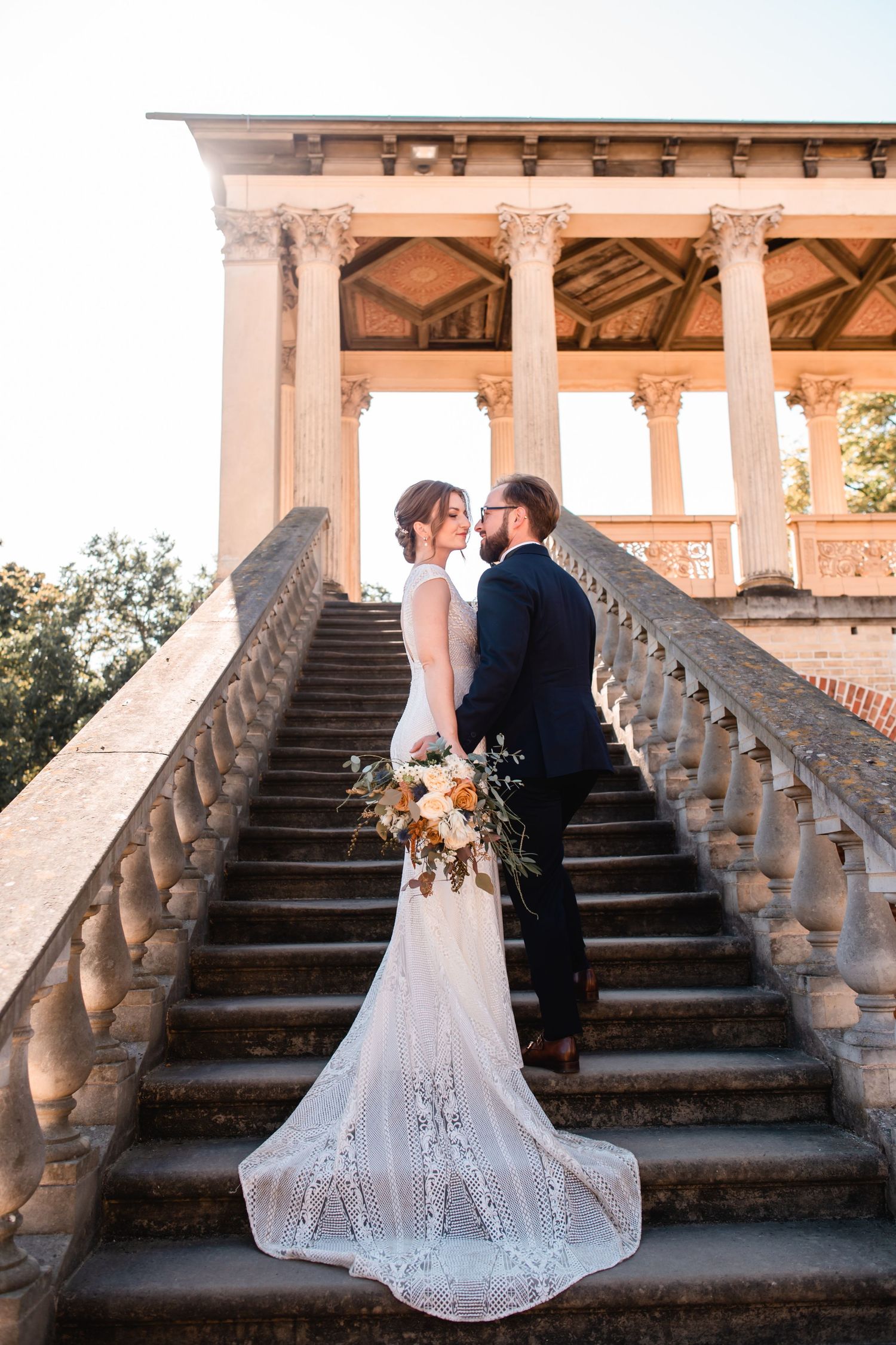 A couple in wedding attire pose on grand stone steps with ornate balustrades at sunset, the bride's delicate lace train trailing behind.