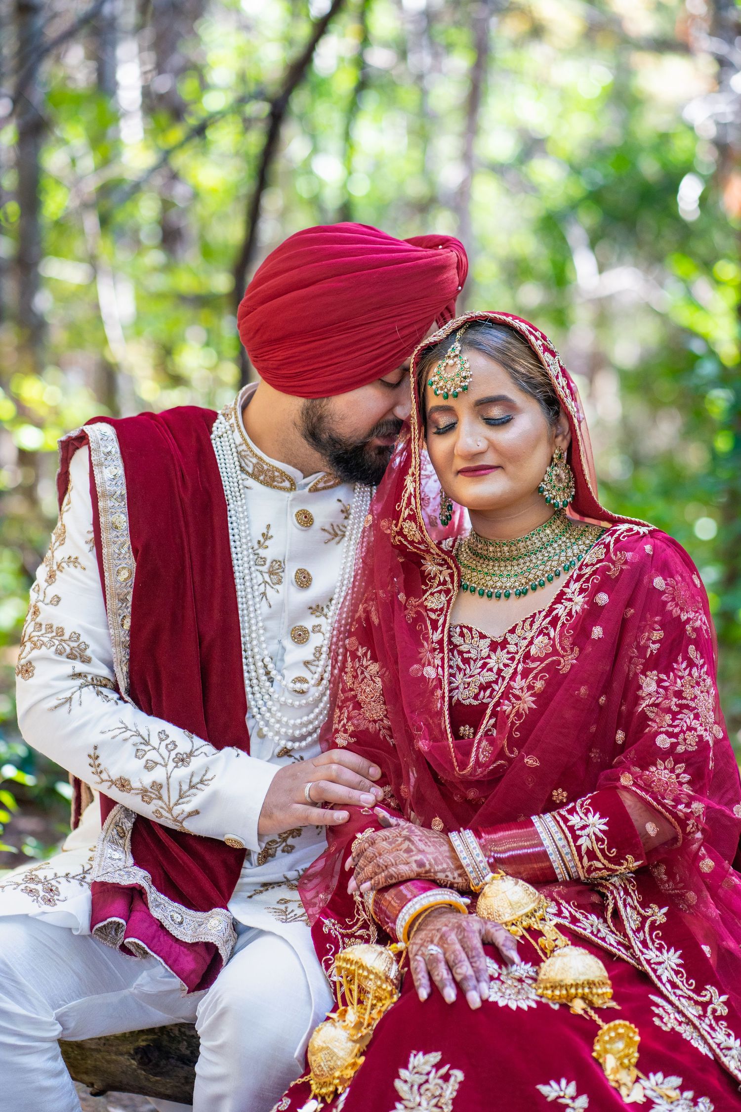 Traditional Sikh wedding couple in red and white attire posing in a garden setting.